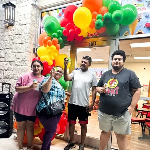 A family raising ice cream cups under the balloon arch outside the shop